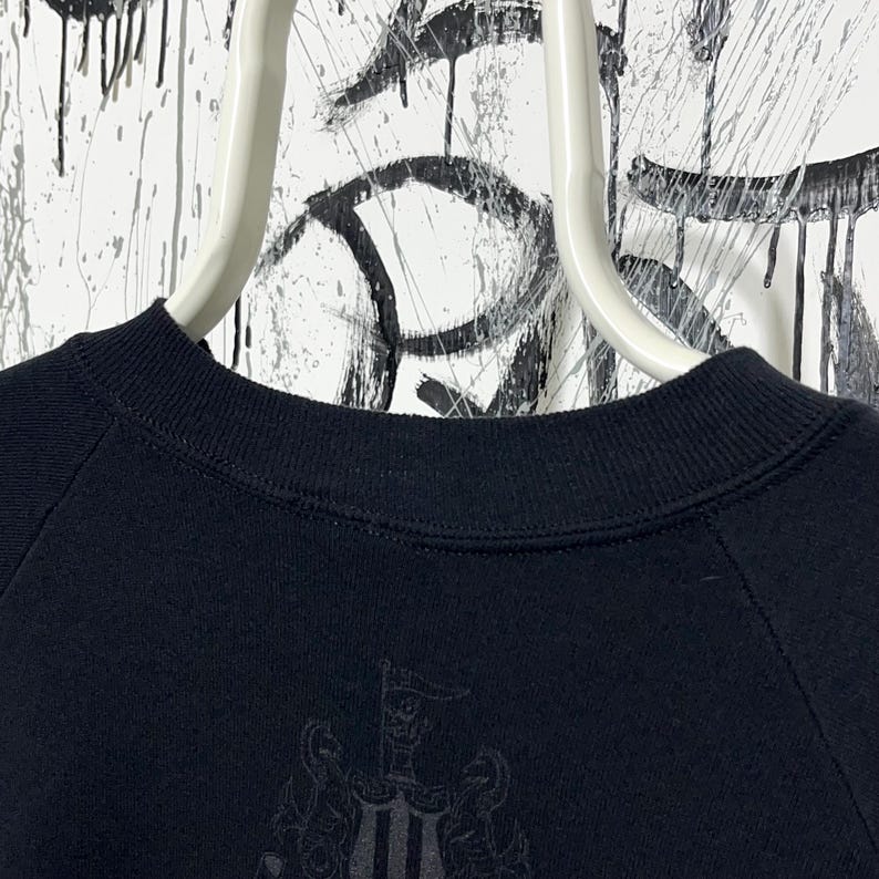 May include: Black crew neck sweatshirt with a tonal emblem on the lower front. The sweatshirt is hanging on a white hanger against a backdrop of black and white abstract art. The emblem appears to be a coat of arms.
