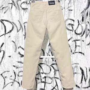 Może przedstawiać: Beżowe dżinsy z klasycznym pięciokieszeniowym designem. Dżinsy mają prosty krój nogawek i wysoki stan. Tylne kieszenie mają subtelne logo. Dżinsy są prezentowane na tle pokrytym graffiti.