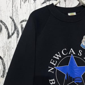 May include: Black crew neck sweatshirt featuring a blue star with the word "NEWCASTLE" in a circular design. The sweatshirt also has a silhouette of a bridge and buildings. The garment is hanging on a white hanger.