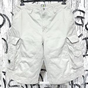 Peut inclure: Shorts cargo blanc cassé avec plusieurs poches et une fermeture boutonnée. Le short porte le logo Adidas à l'avant et est suspendu à un cintre noir. Le short a une coupe décontractée et est fait d'un tissu durable.