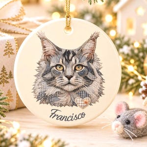 May include: A round white ceramic Christmas ornament featuring a detailed illustration of a grey tabby cat with tufted ears. Below the cat, the name Francisco is printed in black script, and a small heart-shaped patch is visible.