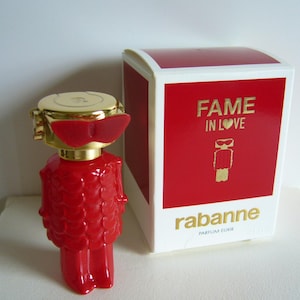 May include: A red perfume bottle with a gold cap and red sunglasses-like accents, next to a red and white box. The box reads "FAME IN LOVE" and "rabanne PARFUM ELIXIR" in gold lettering. The bottle has a unique, textured design.