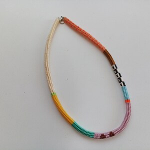 May include: A colorful beaded necklace with a variety of colors including cream, yellow, teal, pink, and brown. The necklace is made of small, round beads strung together to create a unique and eye-catching accessory.