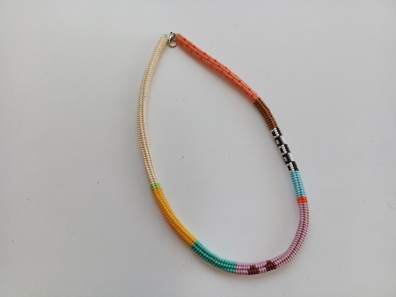 May include: A colorful beaded necklace with a variety of colors including cream, yellow, teal, pink, and brown. The necklace is made of small, round beads strung together to create a unique and eye-catching accessory.