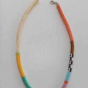 May include: A beaded necklace with a variety of colors. The necklace is made of small beads in sections of cream, yellow, teal, pink, brown, black, white, blue, orange, and coral. It has a silver clasp.