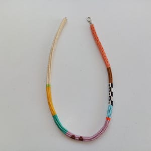 May include: A colorful beaded necklace with a variety of colors including cream, yellow, teal, pink, brown, black, and orange. The necklace has a silver clasp and is made of small, tightly strung beads. The design features a striped pattern.