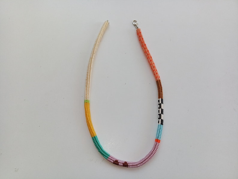 May include: A colorful beaded necklace with a variety of colors including cream, yellow, teal, pink, brown, black, and orange. The necklace has a silver clasp and is made of small, tightly strung beads. The design features a striped pattern.