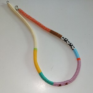 May include: A beaded necklace with a variety of colors. The necklace has a white cord with sections of orange, brown, black and white, light blue, orange, pink, teal, and yellow beads. The necklace is made of small, round beads.