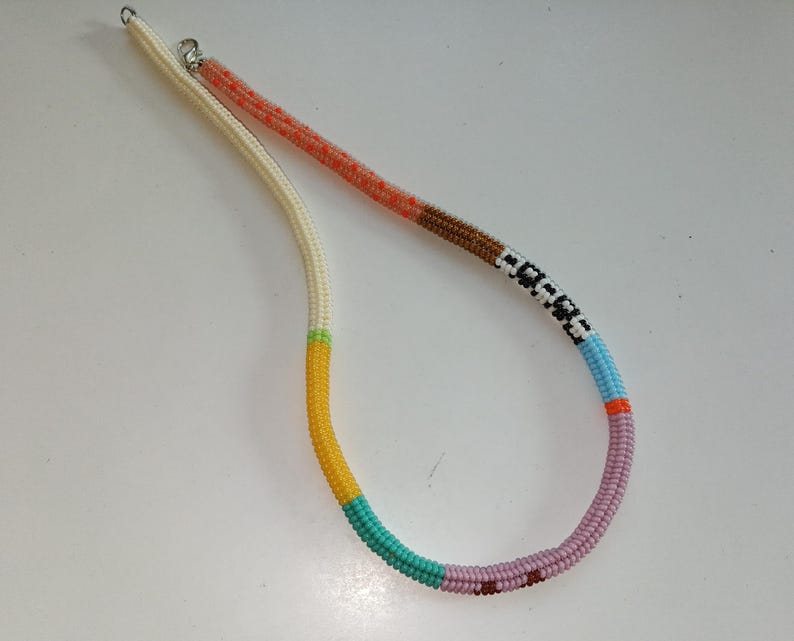May include: A beaded necklace with a variety of colors. The necklace has a white cord with sections of orange, brown, black and white, light blue, orange, pink, teal, and yellow beads. The necklace is made of small, round beads.