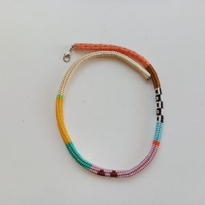 May include: A colorful beaded necklace with a variety of colors including orange, white, yellow, green, turquoise, purple, brown, black, and blue. The necklace has a silver clasp and is laying on a white surface.