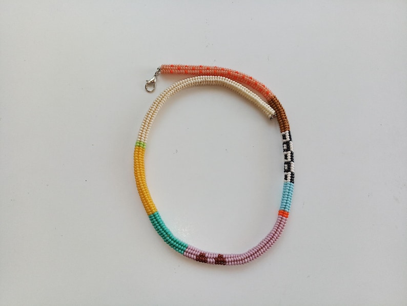 May include: A colorful beaded necklace with a variety of colors including orange, white, yellow, green, turquoise, purple, brown, black, and blue. The necklace has a silver clasp and is laying on a white surface.