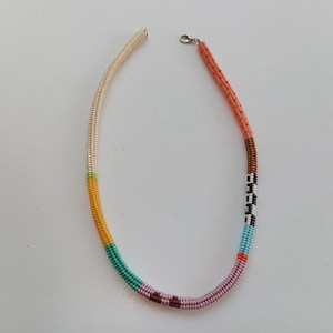 May include: A beaded necklace with a variety of colors. The necklace has sections of cream, yellow, teal, pink, brown, black and white, light blue, and orange beads. The necklace is secured with a silver clasp.