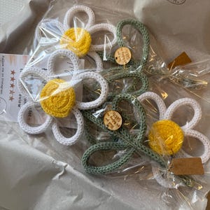May include: Several crocheted flower ornaments packaged in clear plastic. The flowers feature white petals, bright yellow centers, and sage green stems. Small round wooden tags are attached to the stems with the text Thank you for helping me GROW.