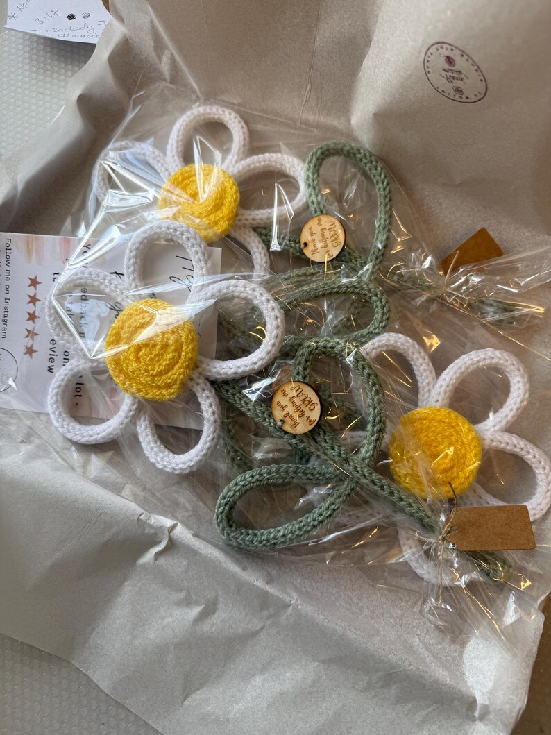 May include: Several crocheted flower ornaments packaged in clear plastic. The flowers feature white petals, bright yellow centers, and sage green stems. Small round wooden tags are attached to the stems with the text Thank you for helping me GROW.