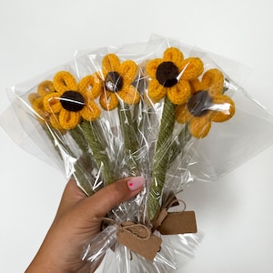 Könnte beinhalten: Ein Strauß aus sechs gehäkelten Sonnenblumen mit braunen Zentren, verpackt in durchsichtigem Plastik und mit Schnur und braunen Papieretiketten gebunden.