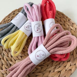 May include: Five bundles of thick, knitted cord in grey, yellow, light pink, dusty rose, and coral. Each bundle is secured with a white paper band featuring the text "Hand made" and "Follow us" with a small yarn ball icon in the centre.