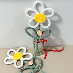 May include: Two crocheted daisy flowers with white petals and yellow centers, featuring green stems and leaves. One flower is placed in a clear glass bottle with a red ribbon, and both have small wooden tags with the text Thank you for helping me grow.
