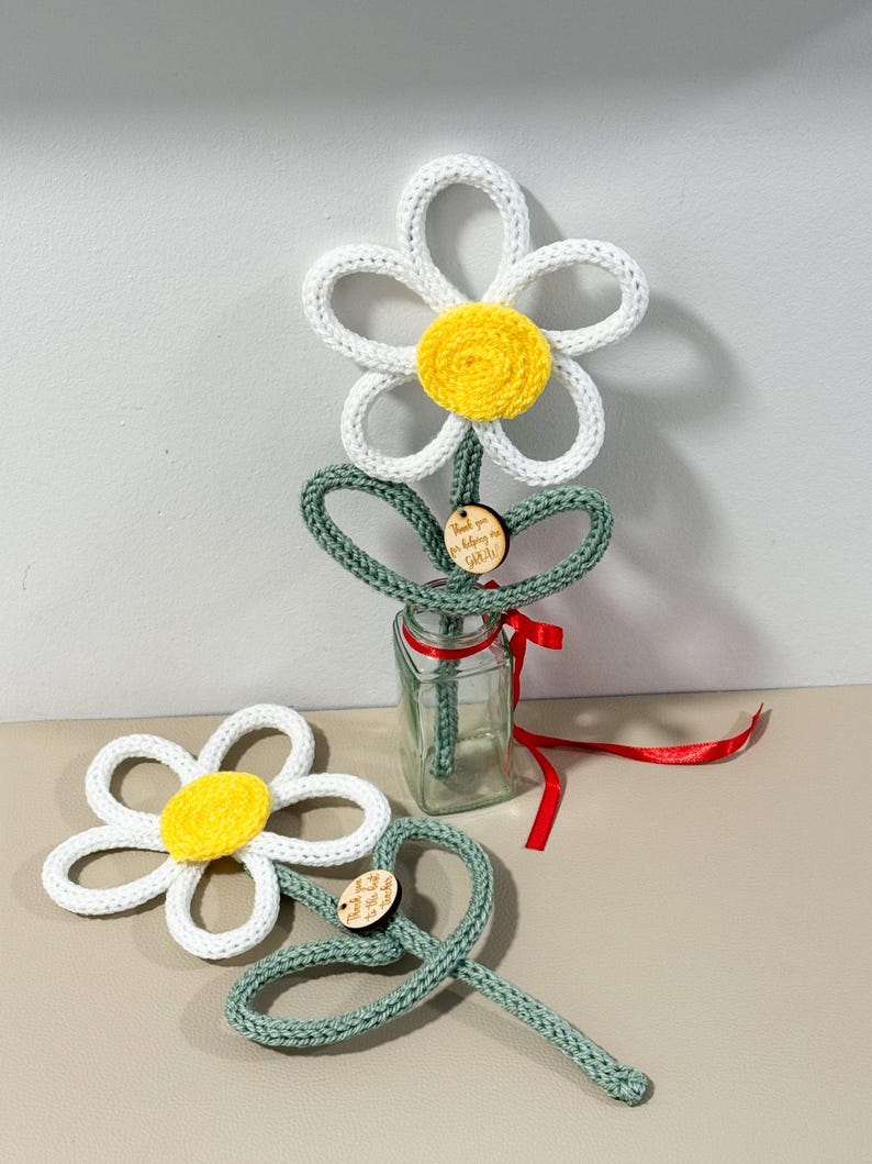 May include: Two crocheted daisy flowers with white petals and yellow centers, featuring green stems and leaves. One flower is placed in a clear glass bottle with a red ribbon, and both have small wooden tags with the text Thank you for helping me grow.