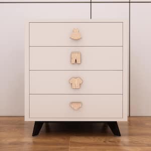 May include: A cream-coloured, four-drawer chest of drawers with black legs. Each drawer has a wooden knob shaped like an item of clothing: a dress, trousers, a shirt, and underwear. The chest is against a neutral background.