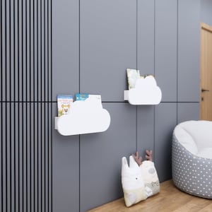 Pode incluir: Prateleiras brancas em forma de nuvem montadas numa parede cinzenta, com livros infantis. Uma cadeira de saco cinzenta e branca com bolinhas e uma almofada decorativa com um desenho de coelho e veado também estão na sala. A sala tem um chão de madeira e uma porta de madeira.