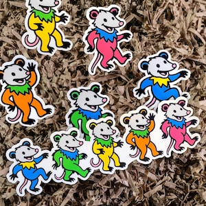 May include: A collection of colorful opossum stickers. Each sticker features a cartoon opossum in various colors, including yellow, pink, orange, green, and blue. The opossums have white bodies, gray heads, and pink tails. The stickers are on a brown, textured background.