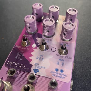 May include: A purple and pink effects pedal with the name "MOOD MKII" printed on the front. The pedal has multiple knobs, switches, and labels, including "SYNC", "LOOP WET", "ENV TAPE STRETCH", and "BOTH FOOT-SWITCHES FOR SECONDARY".