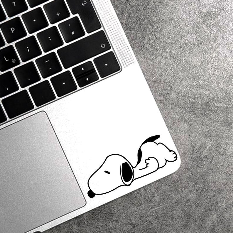 May include: A black and white Snoopy decal is affixed to the corner of a laptop. The decal depicts Snoopy lying down, with his head and front paws visible. The laptop is silver with a black keyboard.