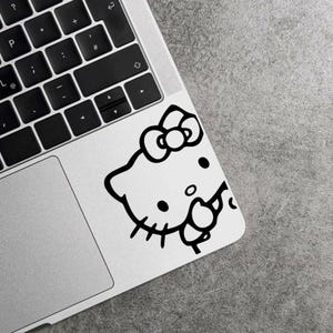 Puede incluir: Una pegatina de Hello Kitty en blanco y negro está pegada en la esquina de un portátil. La pegatina muestra la cara del personaje de dibujos animados, incluyendo su lazo, ojos, nariz, bigotes y boca. El portátil es plateado con un teclado negro.