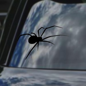 May include: A black spider silhouette decal, with eight legs and a rounded body, is affixed to a car window. The background shows a blurred view of a cloudy sky, creating a contrast between the dark spider and the lighter backdrop.