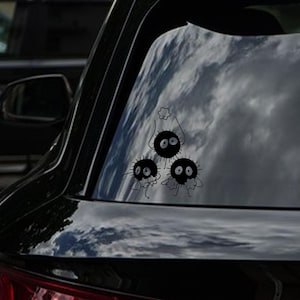 May include: Black vinyl car decal featuring three soot sprites, each with large eyes and small limbs. The top sprite holds a star-shaped object. The decal is applied to a car window, with a cloudy sky reflected in the glass.