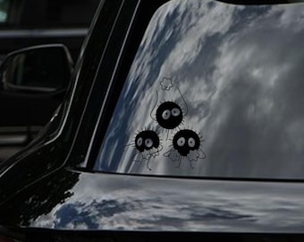 May include: Black vinyl car sticker depicting three soot sprites. Each sprite has large eyes and small limbs. The top sprite holds a star-shaped object. The sticker is applied to a car window, with a cloudy sky reflected in the glass.