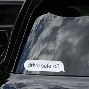 May include: A clear sticker with the text "drive safe <3" in a white speech bubble shape. The sticker is adhered to the rear window of a black vehicle, with reflections of clouds visible.