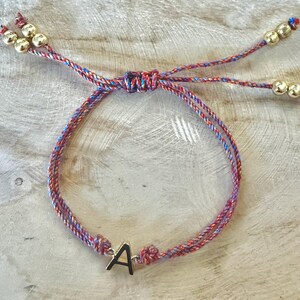 Pode incluir: Pulseira artesanal com cordão trançado em tons de vermelho, azul e roxo. A pulseira apresenta um berloque de metal dourado com a letra "A" e contas douradas nas extremidades das amarrações ajustáveis. A pulseira é exibida em uma superfície de madeira clara.
