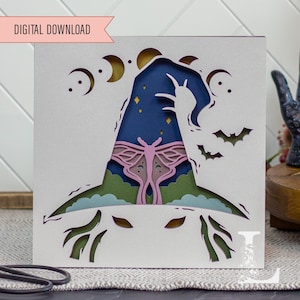 May include: A layered paper art piece with a witch hat design. The hat is filled with a starry night sky, a pink moth, and crescent moons. The words "DIGITAL DOWNLOAD" are in a pink banner at the top.