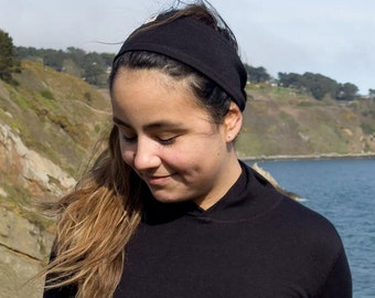 Puede incluir: Una mujer con el pelo largo y castaño lleva una camiseta negra de cuello alto y una diadema negra. Está de pie frente a una masa de agua y un acantilado rocoso.