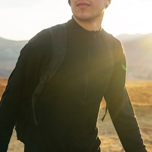 May include: A person wearing a black mock neck shirt with a zipper and a black backpack. The person is standing in a field with mountains in the background.