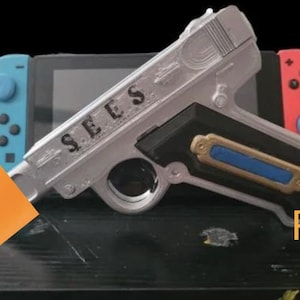 May include: A silver prop toy gun with the word "SEES" printed on the side, resting on a black surface. A Nintendo Switch console is in the background, with blue and red controllers. The word "PROP TOY" is written in orange.
