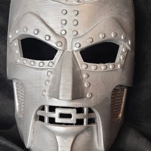 May include: A silver-colored mask with a detailed design, featuring a raised, textured surface and a grid-like mouth opening. The mask has a futuristic, robotic aesthetic.
