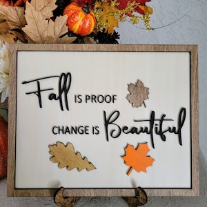 May include: Wooden sign with the text "Fall is proof change is beautiful" in black lettering. The sign has three fall-colored leaves attached to it.