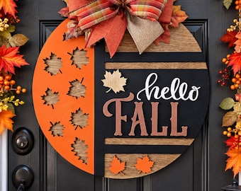 May include: A circular wooden door sign featuring an orange section with cut-out maple leaves and a black section with the text "hello FALL" in white and brown script. The sign is topped with a large decorative bow made of plaid and burlap ribbon.