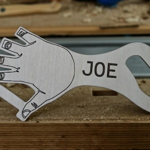 May include: A light-colored wooden bottle opener shaped like a hand, with the name "JOE" printed in black. The opener has a finger-like design and a hole for hanging. It rests on a wooden surface.