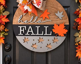 May include: A circular wooden door sign featuring a natural wood top section, a black middle band, and a gray bottom section. The sign displays the text "hello FALL" in white lettering, accented with orange and gray maple leaf cutouts and a large bow.