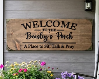 May include: A rectangular wooden sign with dark metal corner accents featuring the black text WELCOME TO THE Beasley's Porch A Place to Sit, Talk & Pray. The sign has a natural wood grain finish and is mounted horizontally on a gray wall.