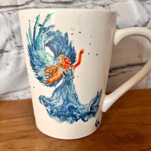 May include: White ceramic mug with a hand-painted design of a woman with flowing blue hair and wings, surrounded by blue and green waves.