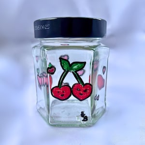 Puede incluir: Un tarro de vidrio transparente con tapa negra, decorado con un diseño pintado a mano de dos cerezas rojas con tallos y hojas verdes. Las cerezas tienen caras sonrientes. El tarro está rodeado de corazones rosas.