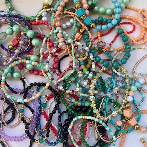 May include: A collection of beaded bracelets in various colors, including green, blue, orange, and black. The bracelets feature different bead sizes and materials, with some incorporating gold-colored accents. The bracelets are scattered on a white surface.