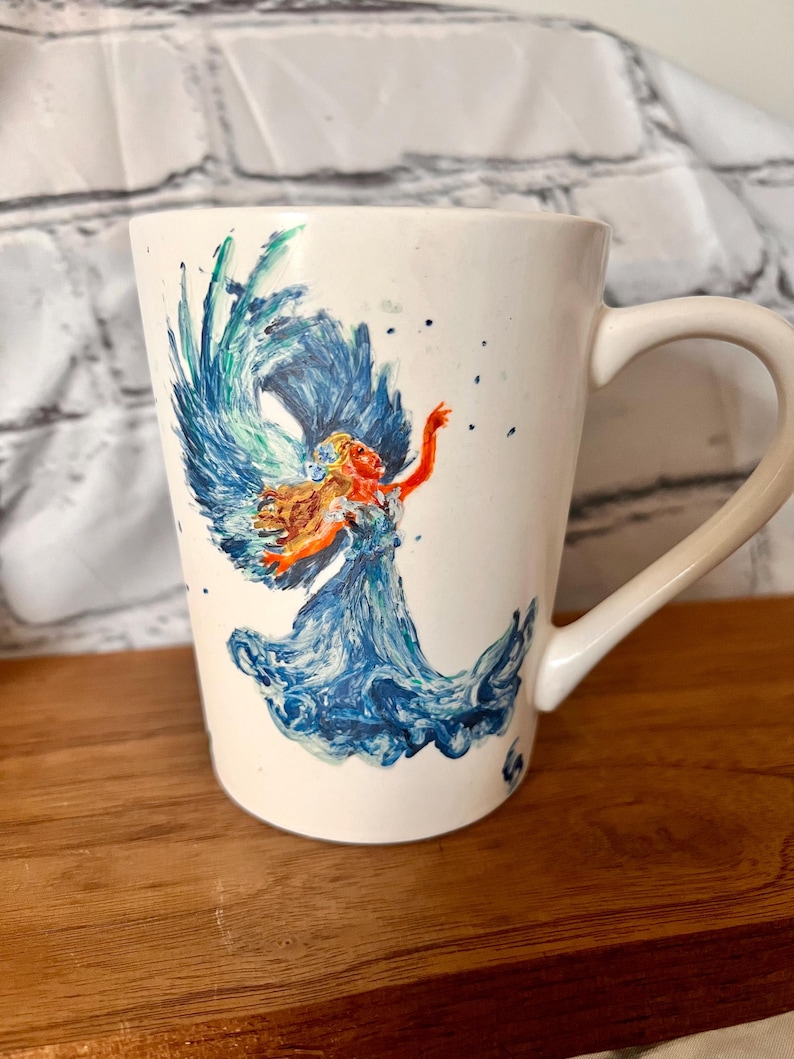 May include: White ceramic mug with a hand-painted design of a woman with flowing blue hair and wings, surrounded by blue and green waves.
