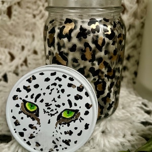 May include: A glass jar with a white lid decorated with a leopard print design in black and gold. The lid features a hand-painted snow leopard face with green eyes.