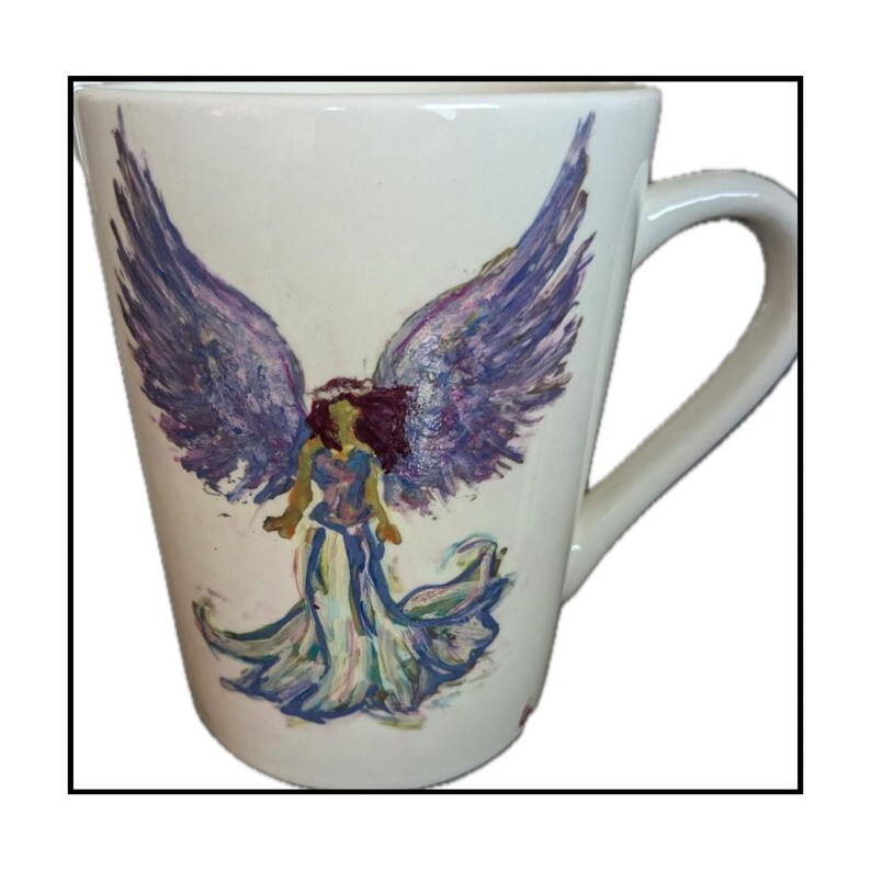 May include: White ceramic mug with a hand-painted design of an angel with purple wings and a flowing gown. The angel is depicted with a purple, blue, and green color scheme.
