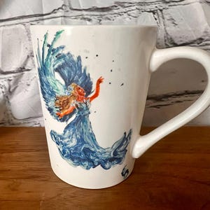 May include: White ceramic mug with a hand-painted design of a woman with blue hair and a blue dress, surrounded by blue and green water-like swirls.